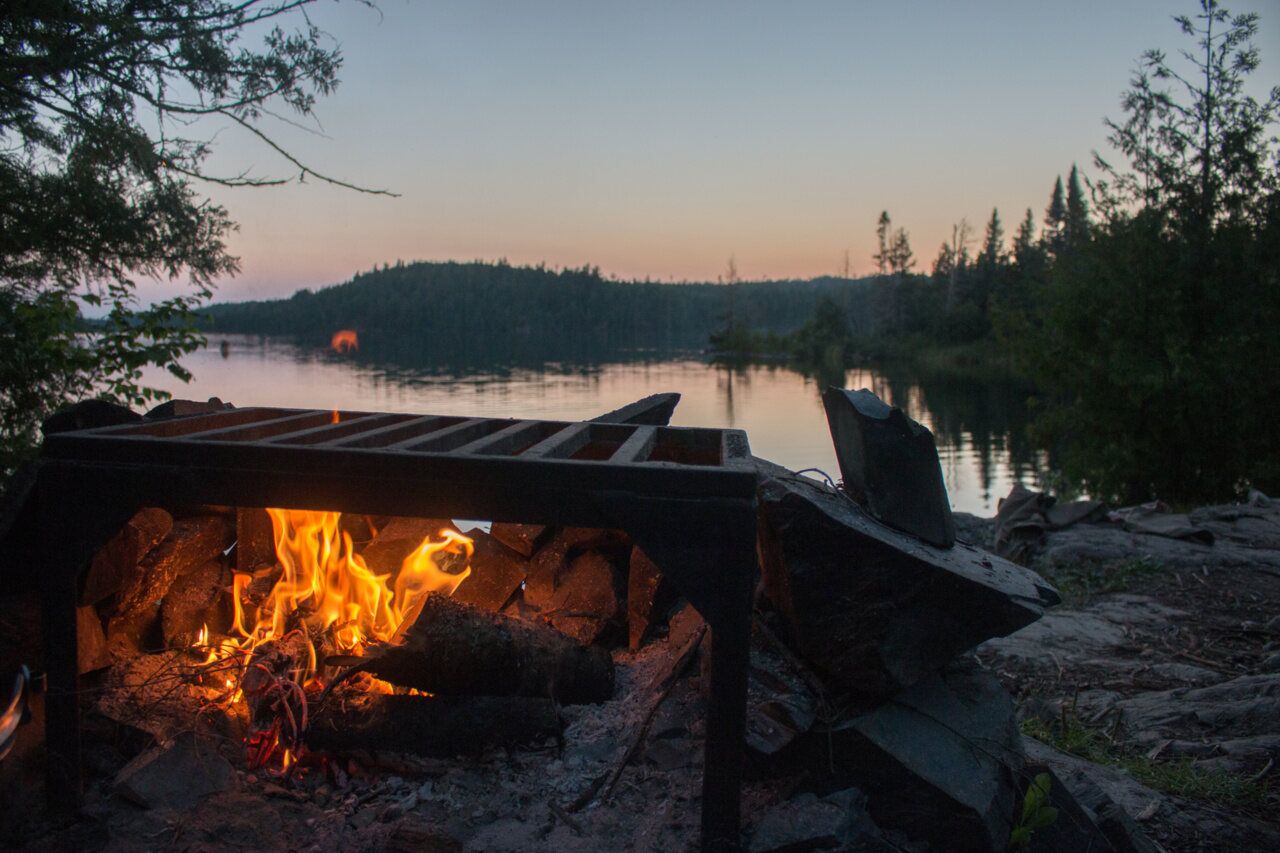 A campfire in an iron grate on a rocky lakeshore at dusk